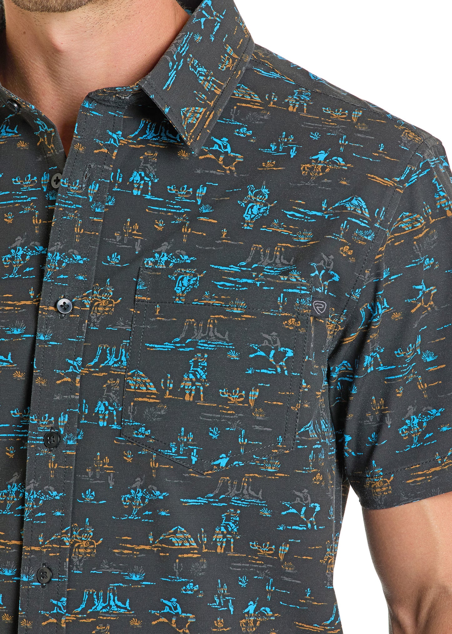 A close-up of a person wearing the Rock & Roll Denim TEK Western Desert Blues Short Sleeve Shirt, a dark UPF 50 shirt with a blue and brown desert and cactus print; only the upper torso is shown.