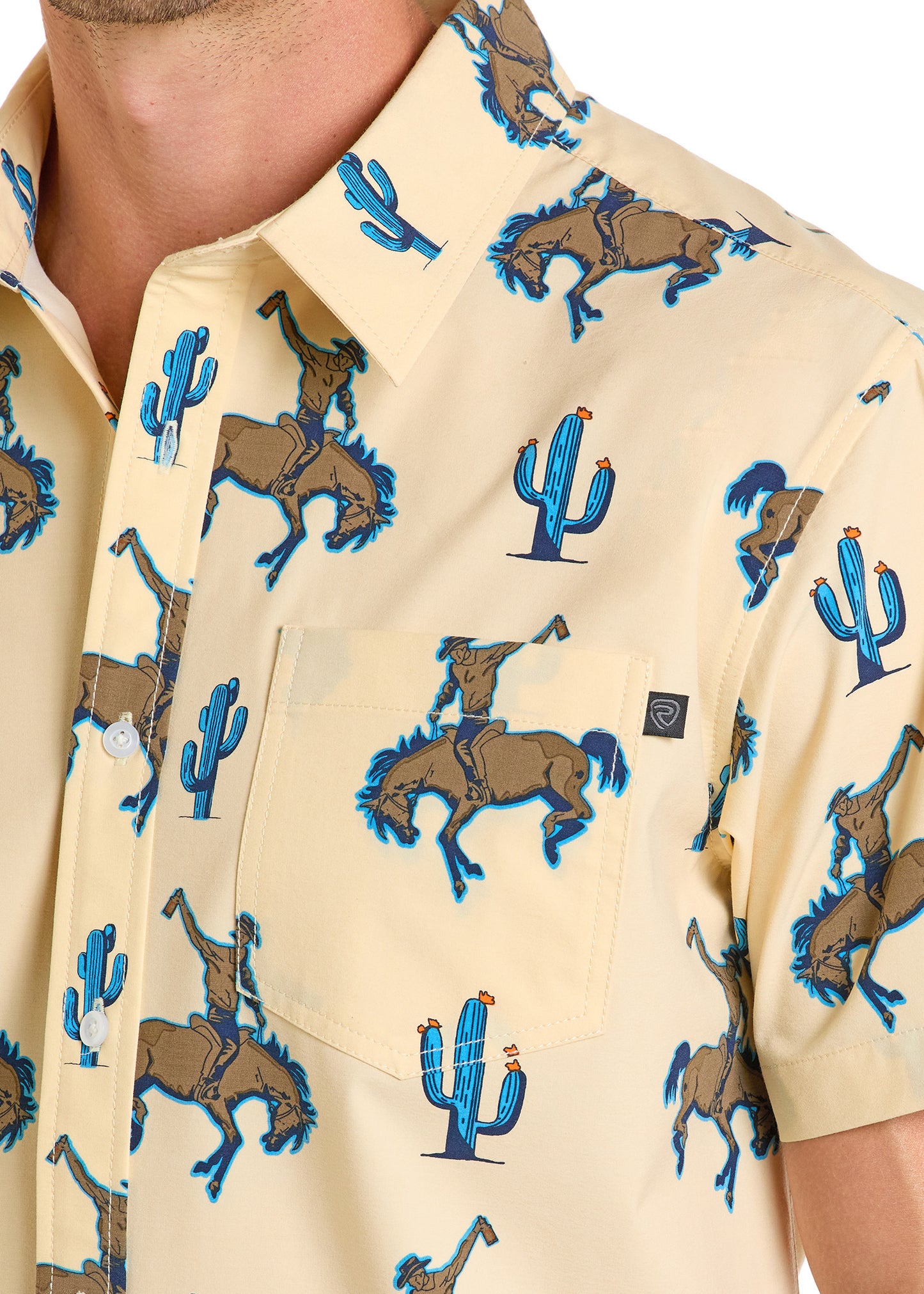 A close-up of the Rock & Roll Denim TEK Western Desert Bronco Short Sleeve Shirt—a beige UPF 50 button-up with blue-outlined cowboys, bucking broncos, cartoon cacti, and a left chest pocket.