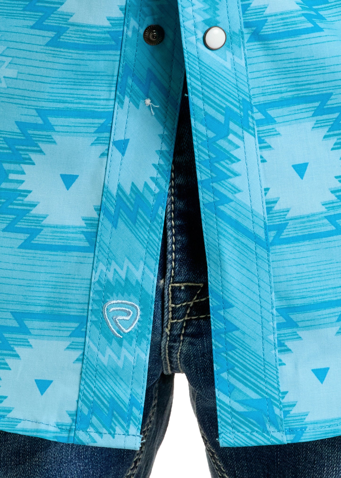 Close-up of the Rock & Roll Denim Slim Fit Turquoise Aztec Long Sleeve Snap Shirt, featuring blue geometric patterns and a small embroidered logo on the hem, worn partially unbuttoned over dark blue jeans.