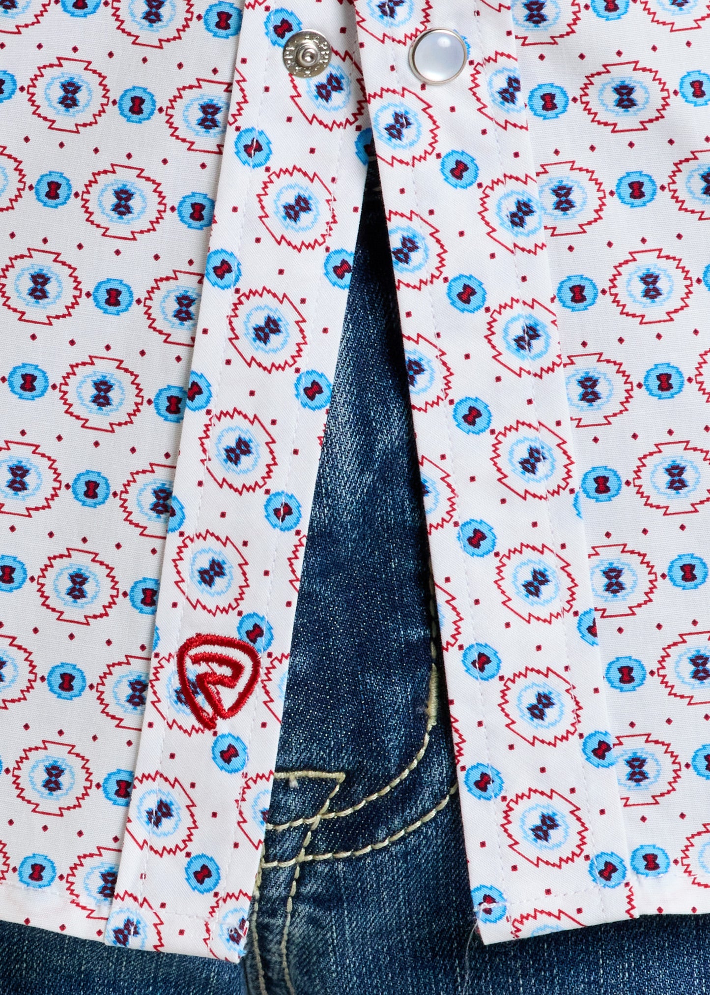 Close-up of Rock & Roll Denim's Slim Fit American Aztec Long Sleeve Snap Shirt with blue and red geometric pattern, partially unbuttoned to show blue denim jeans. A small red embroidered detail appears near the hem.