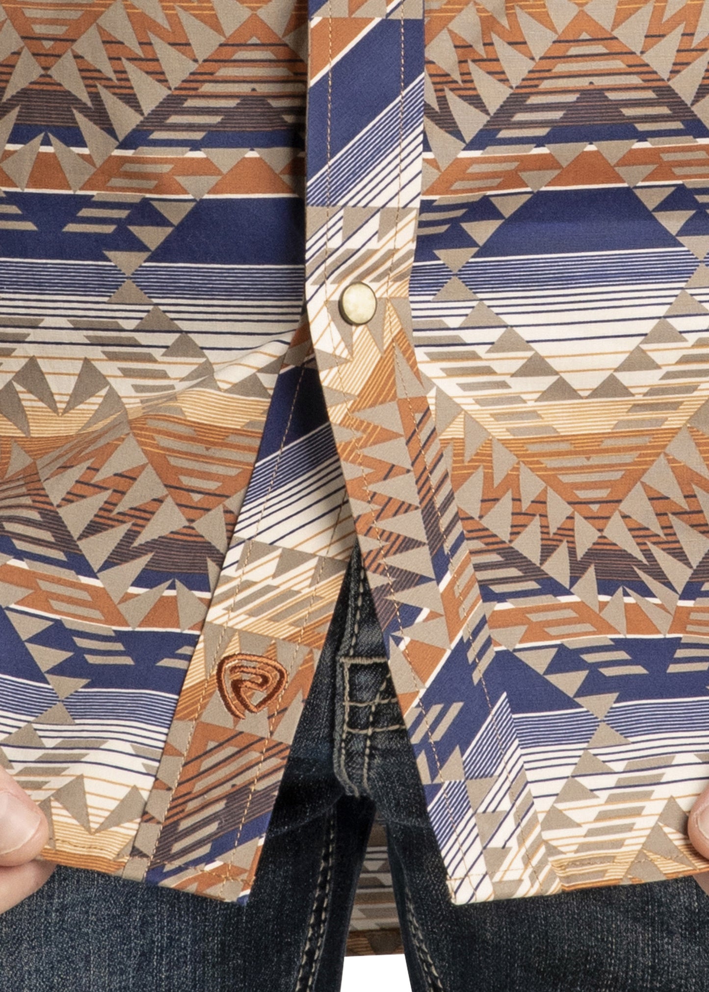 A close-up of a person wearing the Rock & Roll Denim Slim Fit Tan and Blue Striped Aztec Long Sleeve Snap Shirt with blue jeans, holding the sides of the shirt near the bottom button.