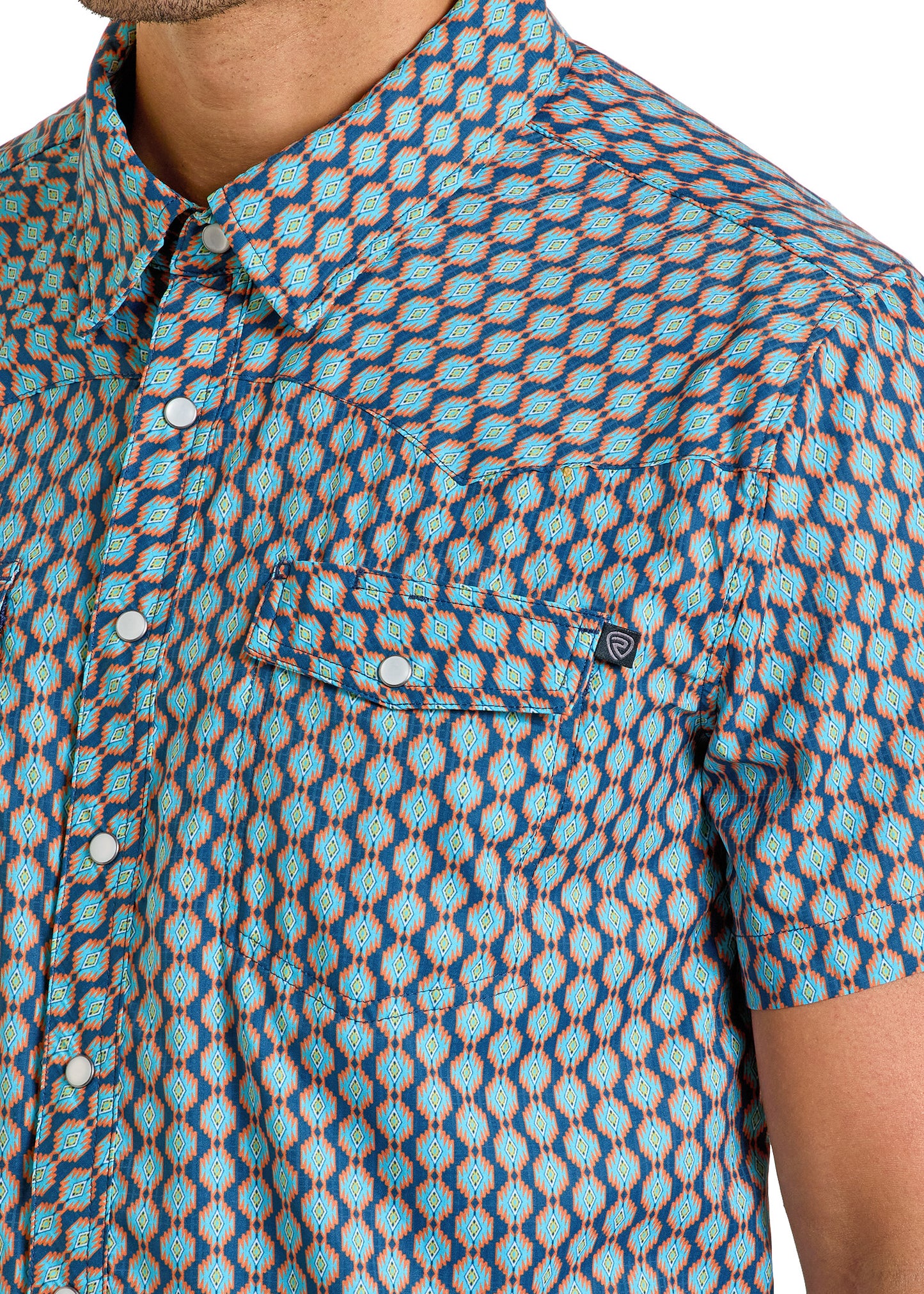 A man wears the Rock & Roll Denim TEK Western Mini Aztec Print Short Sleeve Shirt, featuring blue, teal, and yellow geometric designs, white buttons, and a flap chest pocket with a logo tag. Only his upper torso is visible.