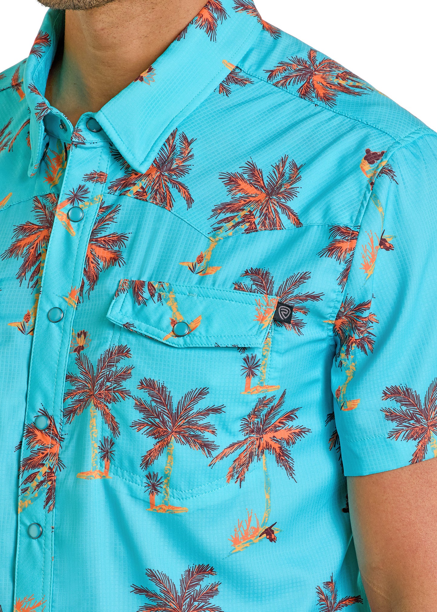 A close-up of a person wearing the Rock & Roll Denim TEK Western Tropical Western Short Sleeve Shirt, featuring turquoise fabric with an orange palm tree pattern, a collar, moisture-wicking material, and a buttoned chest pocket. Only the upper torso is visible.