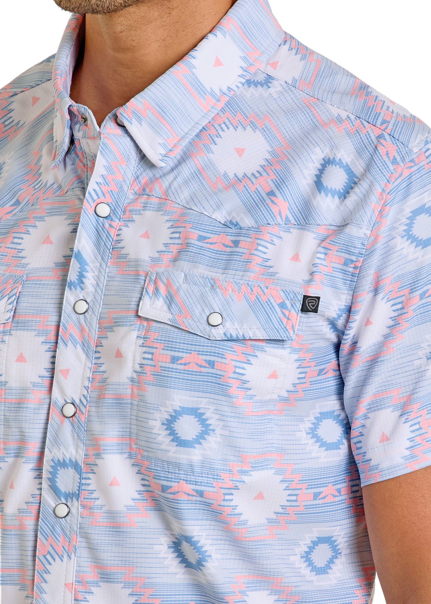 A man wears the TEK Western Light Aztec Short Sleeve Shirt by Rock & Roll Denim in light blue with pink/blue geometric patterns and a chest pocket with logo tag. Only his upper body is visible. The shirt provides moisture-wicking comfort all day.