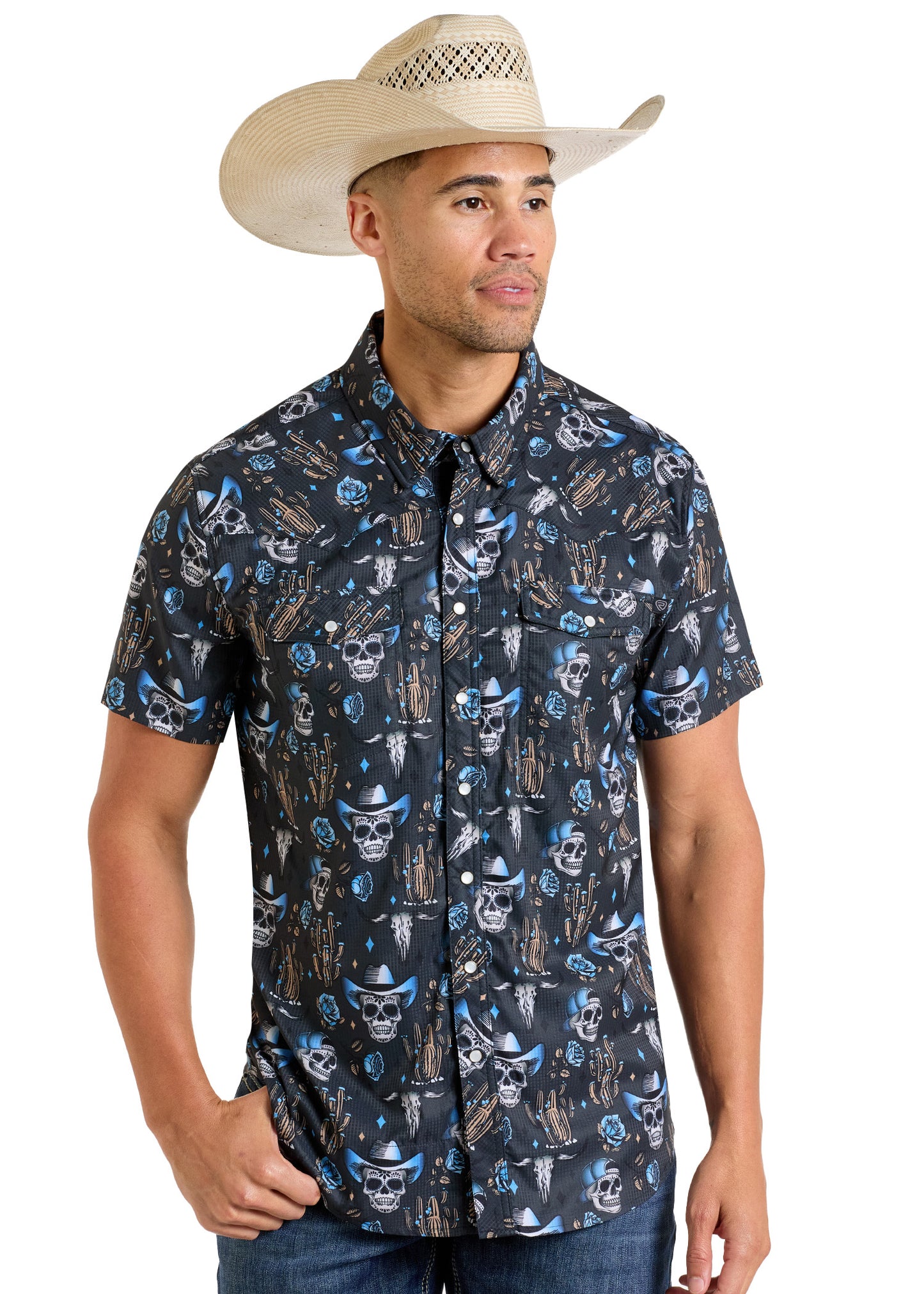 A man wearing a cowboy hat and the Rock & Roll Denim TEK Western Skull Short Sleeve Shirt, featuring black fabric with blue and white patterns, stands with one hand in his pocket, looking slightly to the side.