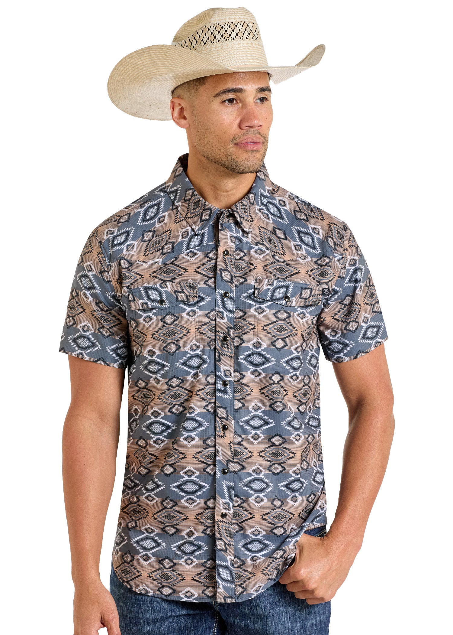 A man wearing a cream cowboy hat and a Rock & Roll Denim TEK Western Bold Aztec Short Sleeve Shirt with tan, blue, and gray geometric patterns stands relaxed, looking slightly to the side.