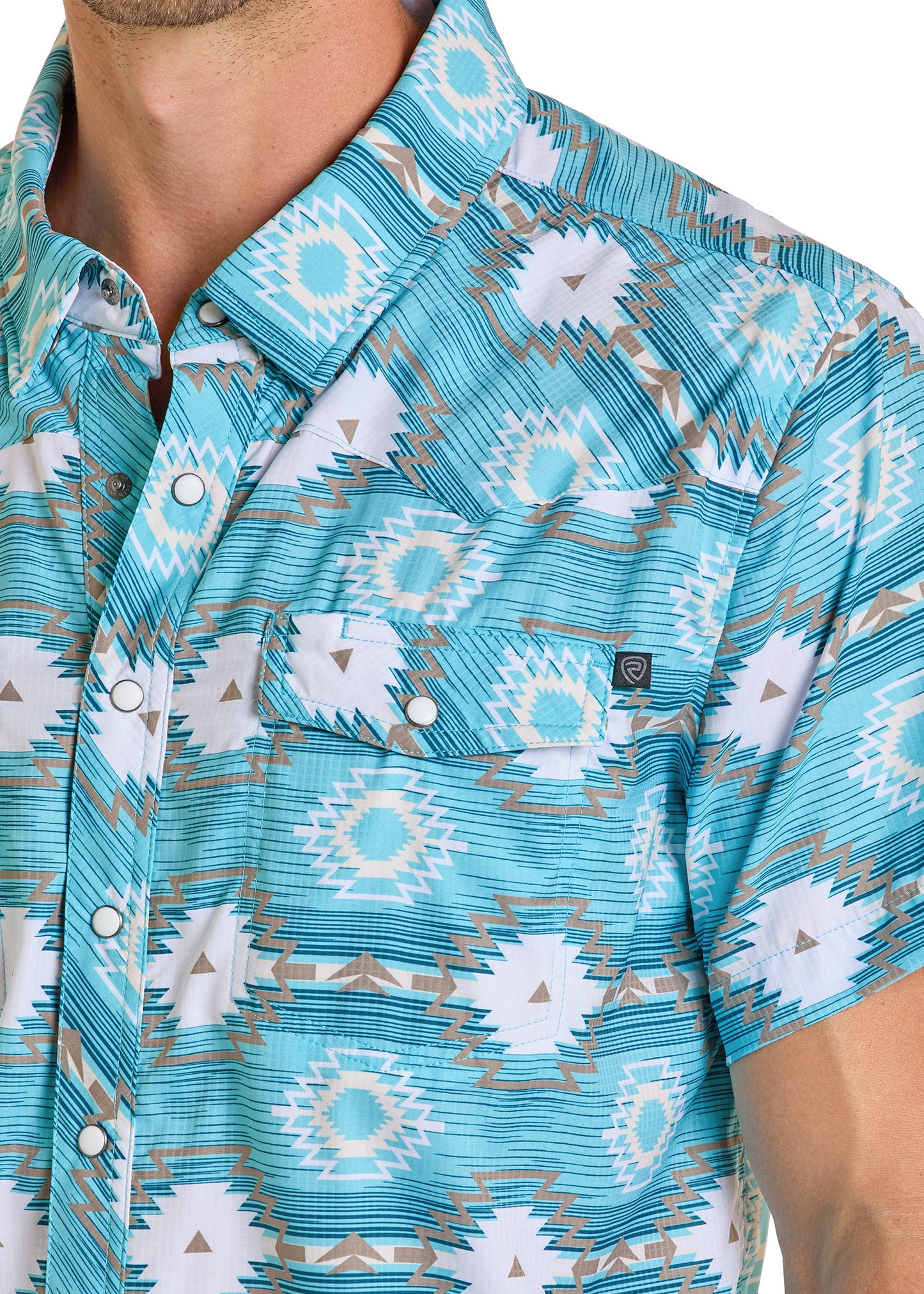 A close-up shows a person wearing the Rock & Roll Denim TEK Western Turquoise Aztec Short Sleeve Shirt with white, gray, and light brown geometric patterns, a front pocket logo, metal snap buttons, and only part of their face visible.