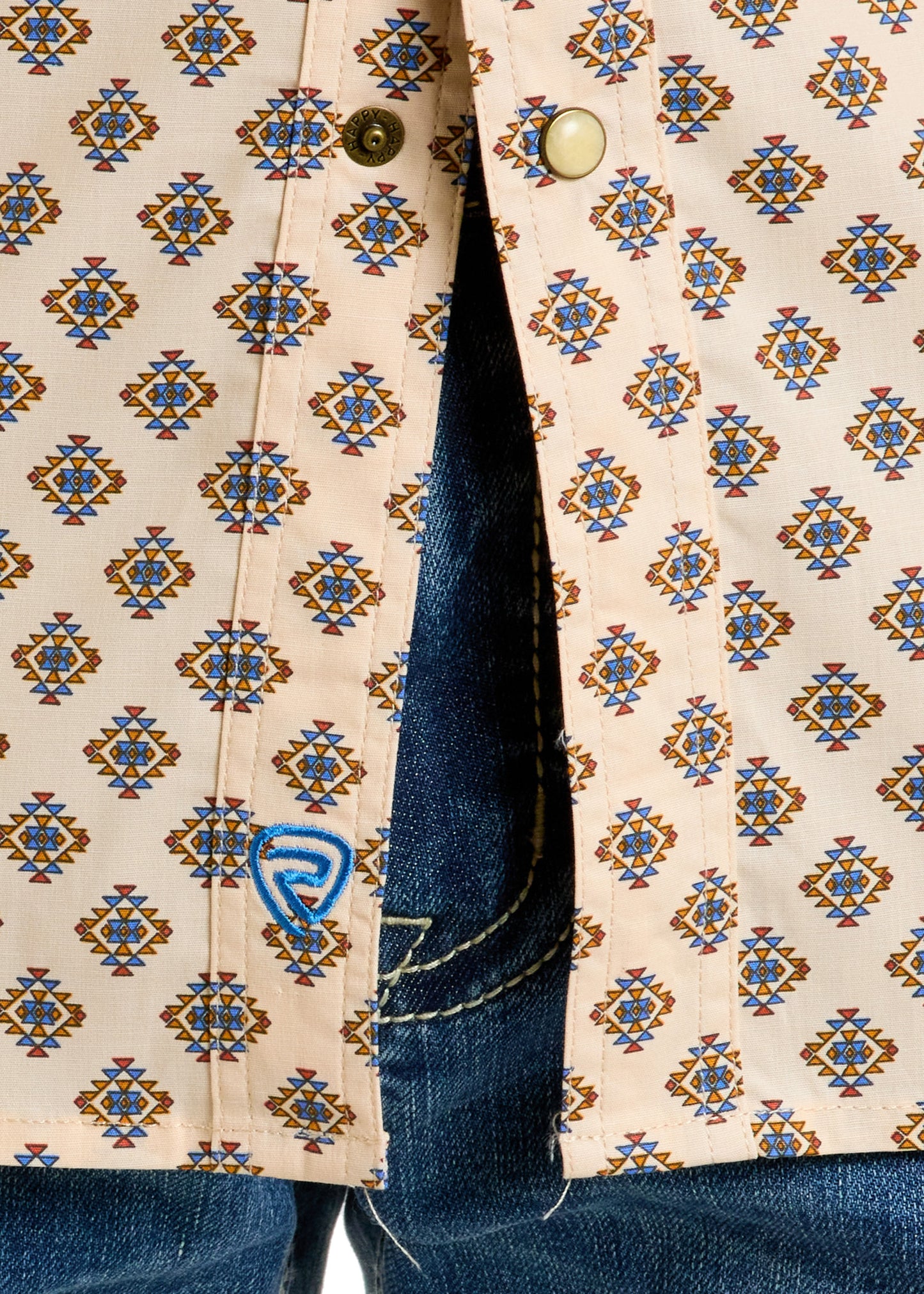 Close-up of the Rock & Roll Denim Slim Fit Geo Aztec Print Short Sleeve Snap Shirt with blue and yellow geometric pattern, gold snaps, blue logo embroidery by the hem, paired with blue jeans showing visible zipper and stitching.