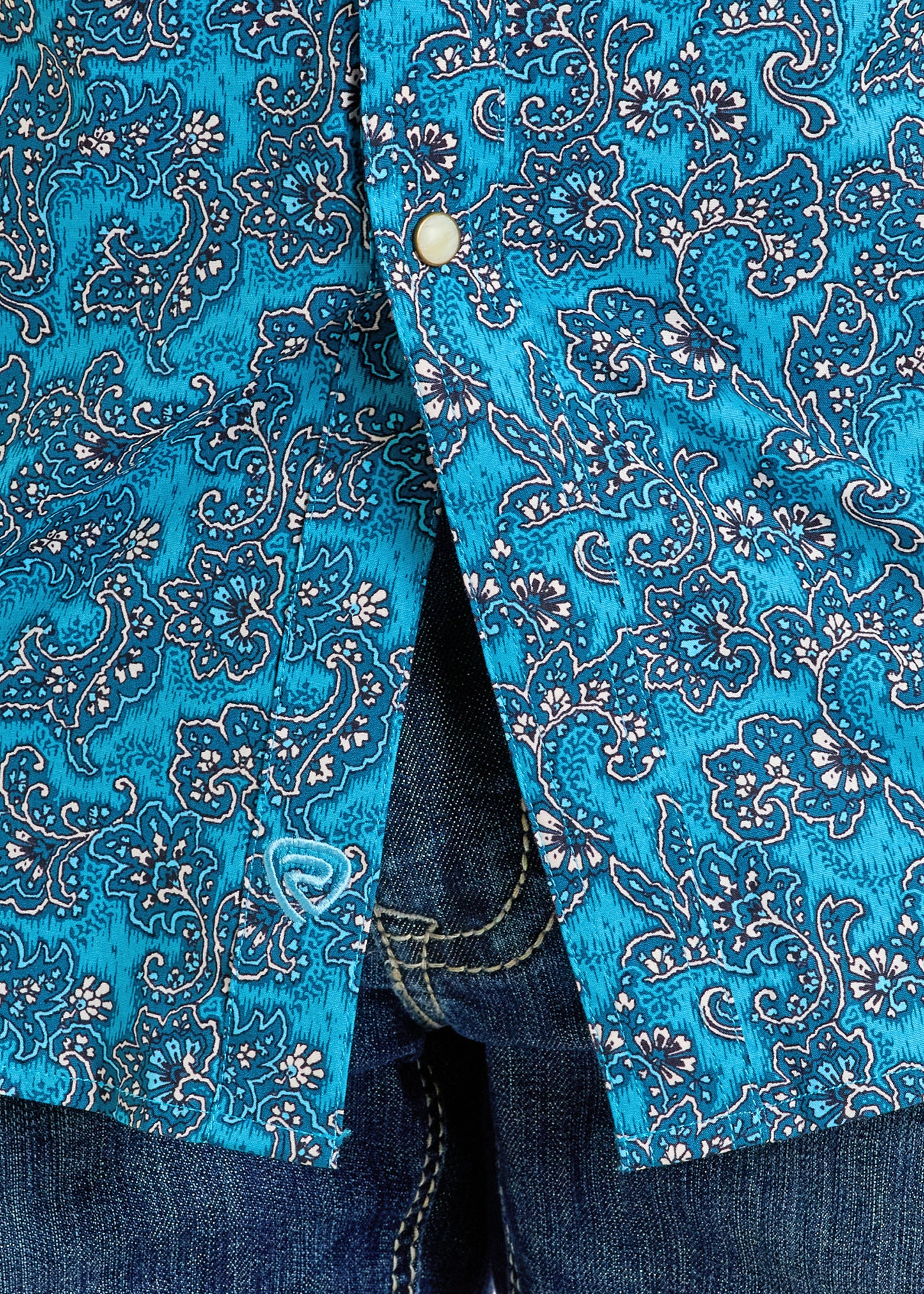 Close-up of the Rock & Roll Denim Slim Fit Turquoise Paisley Short Sleeve Snap Shirt with pearl buttons, partially unbuttoned to reveal blue denim jeans underneath.