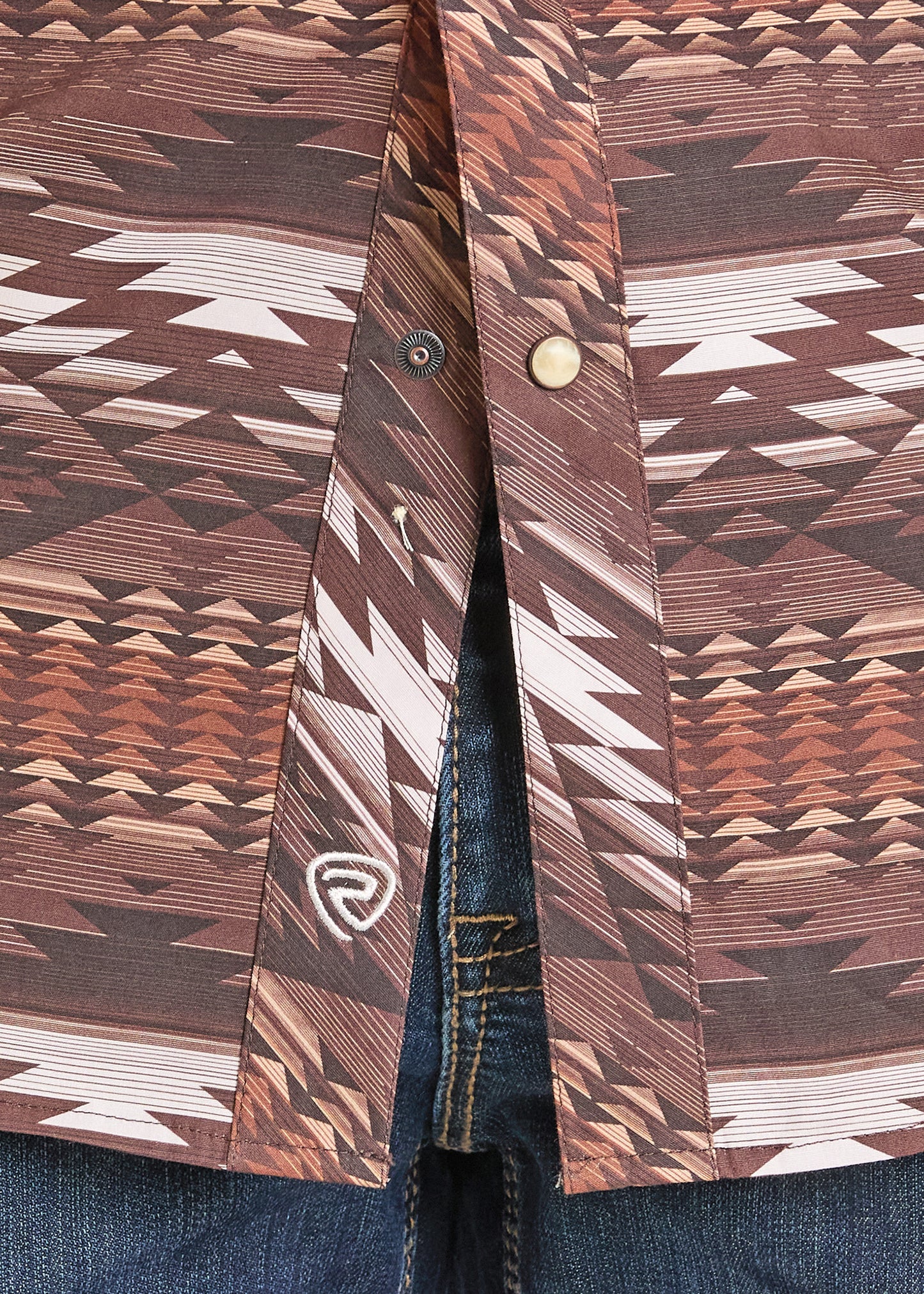 Close-up of the Rock & Roll Denim Slim Fit Brown Aztec Short Sleeve Snap Shirt in size S, with geometric patterns and partially unbuttoned to show the zipper and waistband of blue jeans underneath.