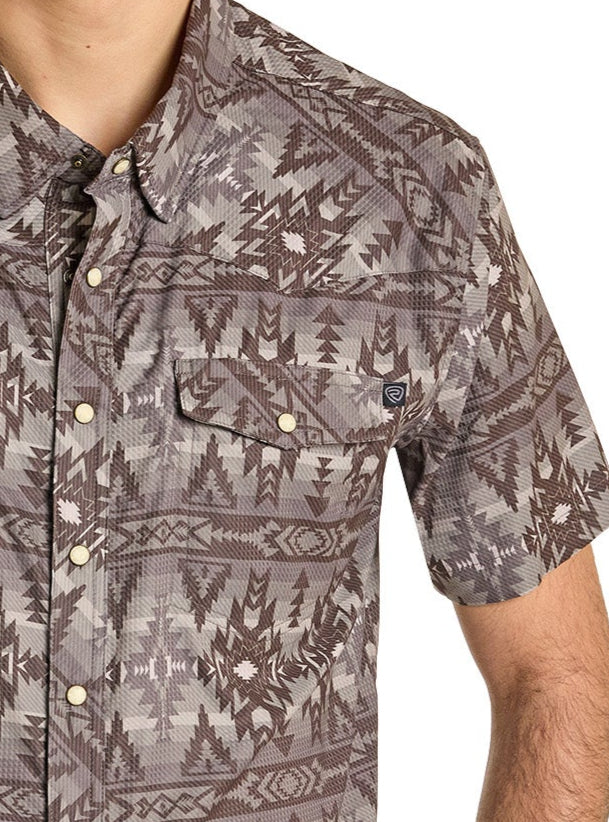 A person is wearing the Rock & Roll Denim TEK Western Brown Aztec Short Sleeve Shirt, featuring a geometric brown and gray pattern and moisture-wicking fabric. Only their upper torso and part of their left arm are visible.