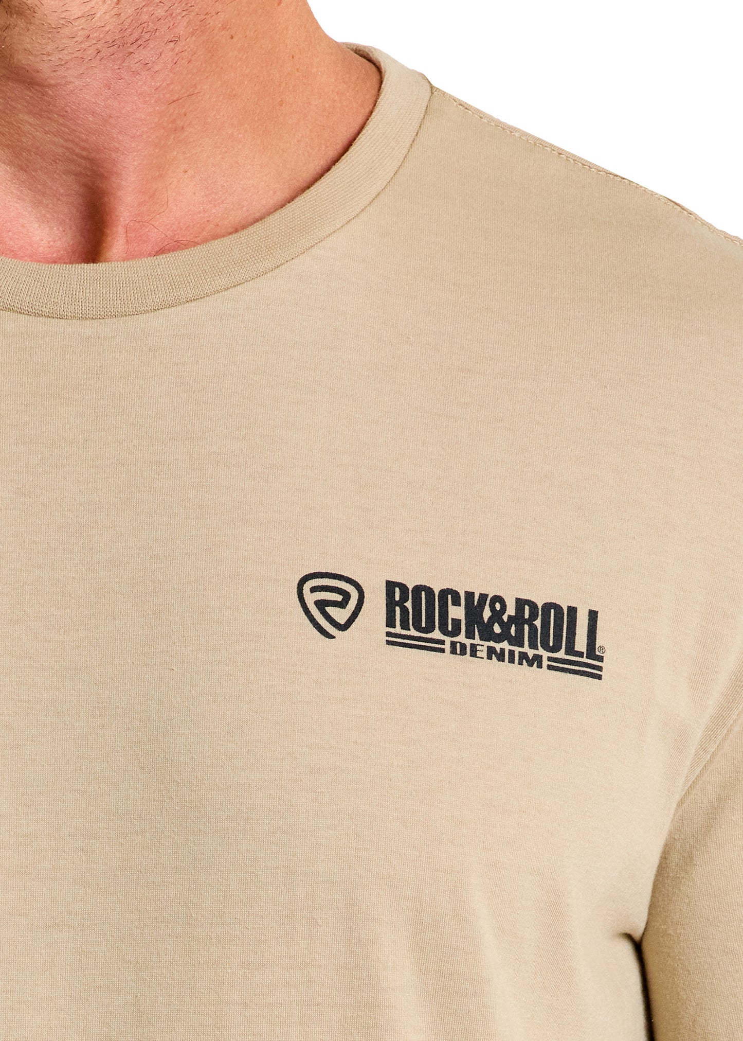 A close-up shows someone wearing the beige Dale Brisby Radiator Ranch Graphic Tee by Rock & Roll Denim, featuring the brand’s logo in black on the left chest. Only the lower face, neck, and upper chest are visible.