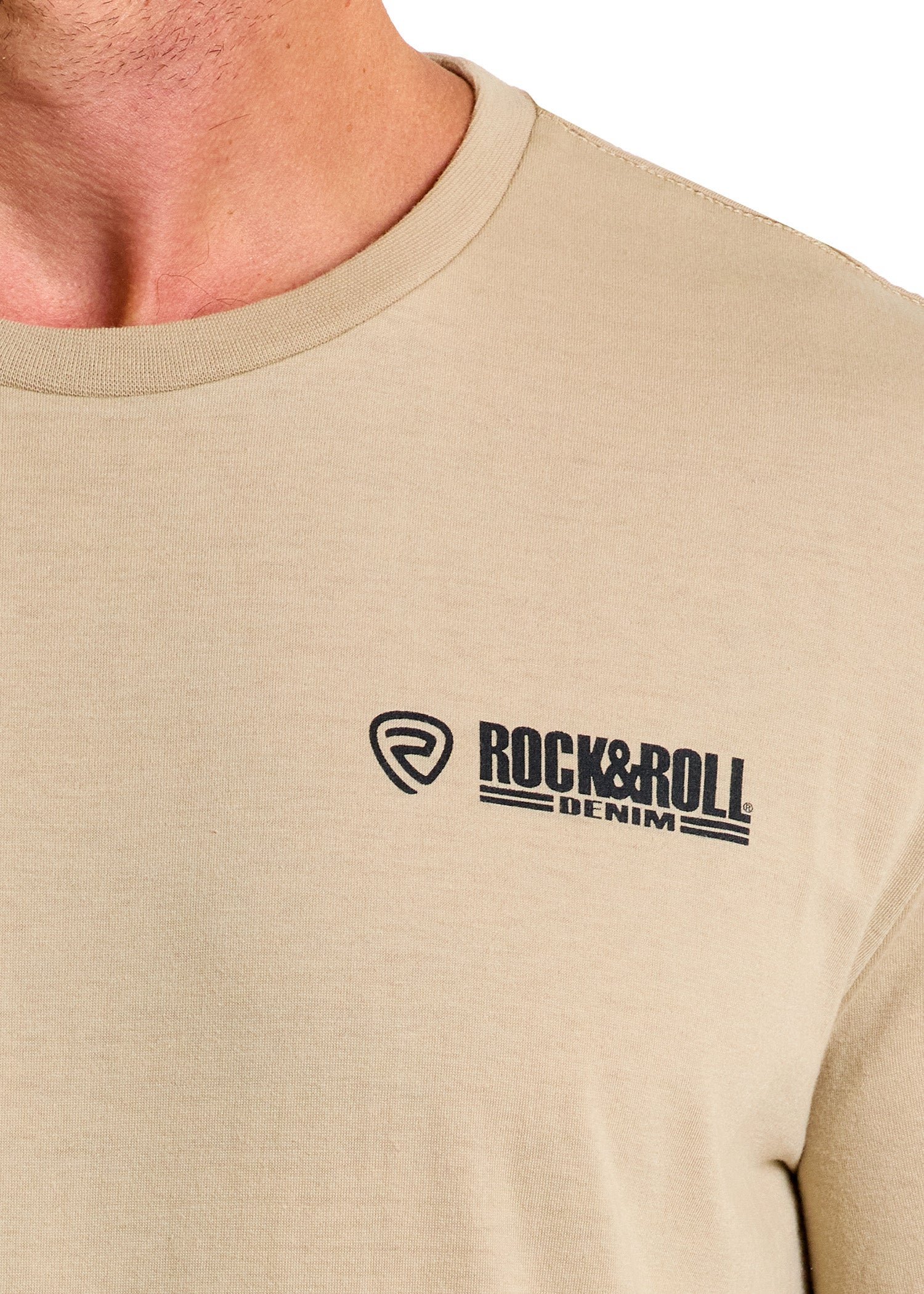 A close-up shows someone wearing the beige Dale Brisby Radiator Ranch Graphic Tee by Rock & Roll Denim, featuring the brand’s logo in black on the left chest. Only the lower face, neck, and upper chest are visible.