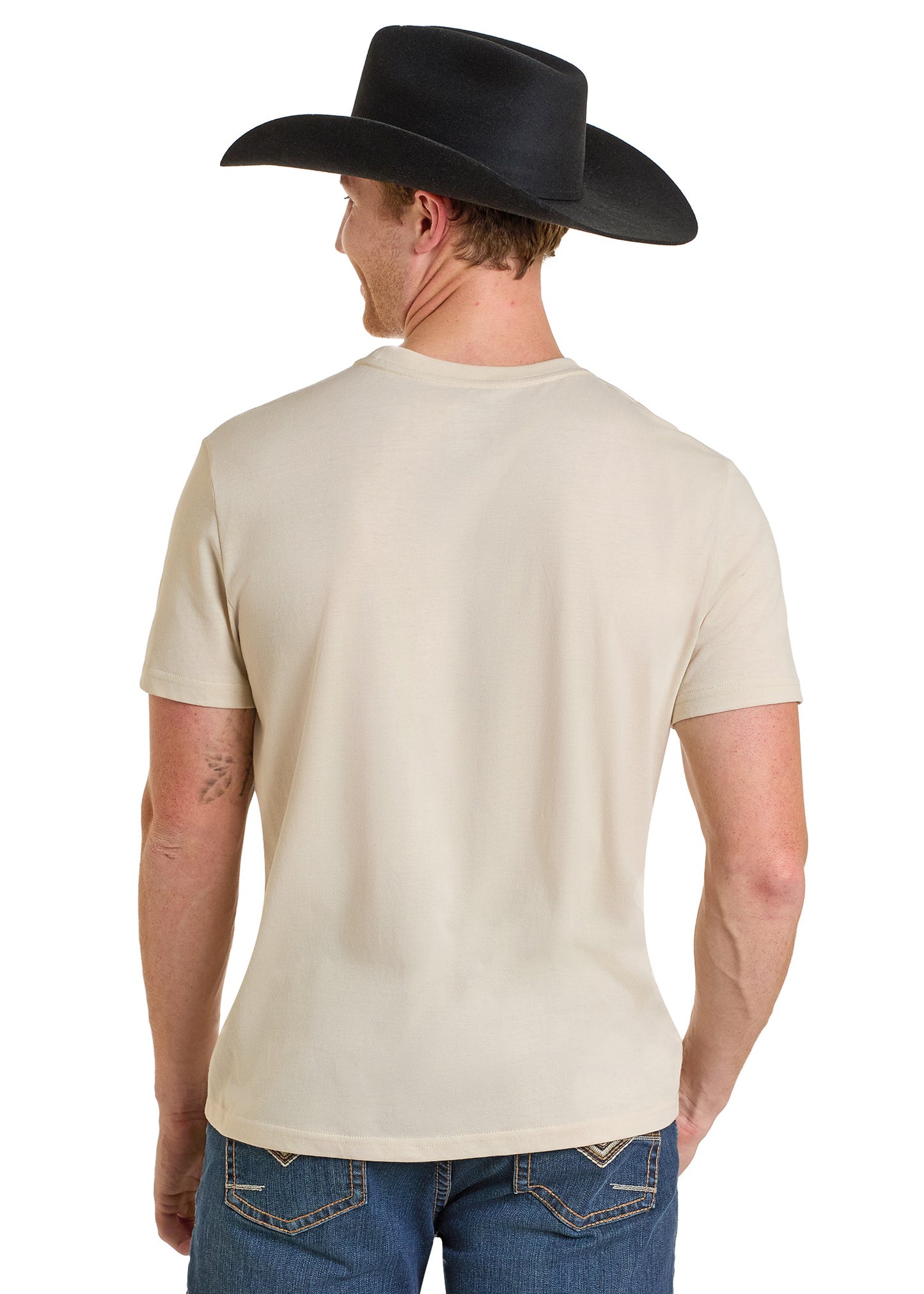 A man wearing a black cowboy hat, blue jeans, and a Rock & Roll Denim Dale Brisby Pow Pow Graphic Tee stands with his back to the camera, displaying the back of his outfit.