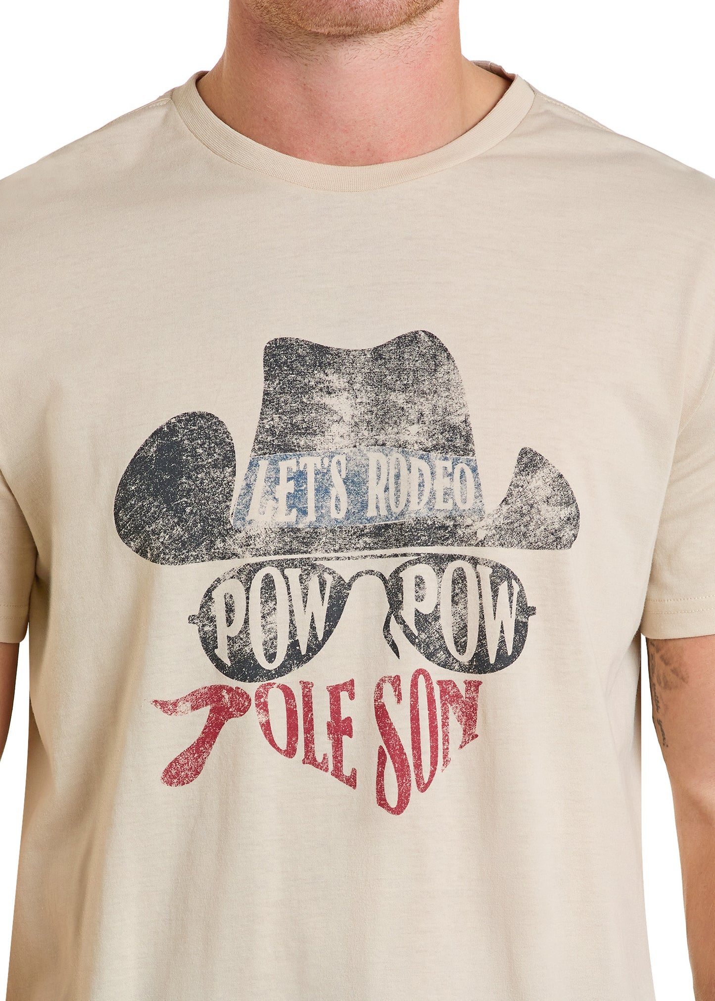 A person wears the Rock & Roll Denim Dale Brisby Pow Pow Graphic Tee—a beige cotton-poly blend featuring a cowboy hat, sunglasses, red bandana, and "LETS RODEO POW POW RODE SOME" graphic.