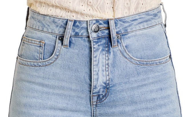 Close-up of a person wearing Rock & Roll Denim’s Mid Rise Distressed Two-Tone Modest Bootcut Jeans with a white, textured, long-sleeved top. The jeans have visible stitching, front button, and zipper fastening.