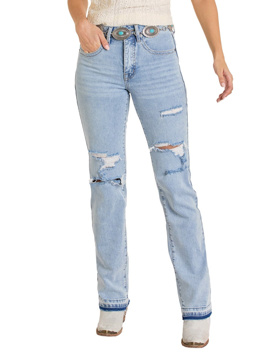 A person models the Rock & Roll Denim Mid Rise Distressed Two-Tone Modest Bootcut Jeans with a decorative belt, white textured top, and white boots—one hand in their pocket and the other relaxed by their side.