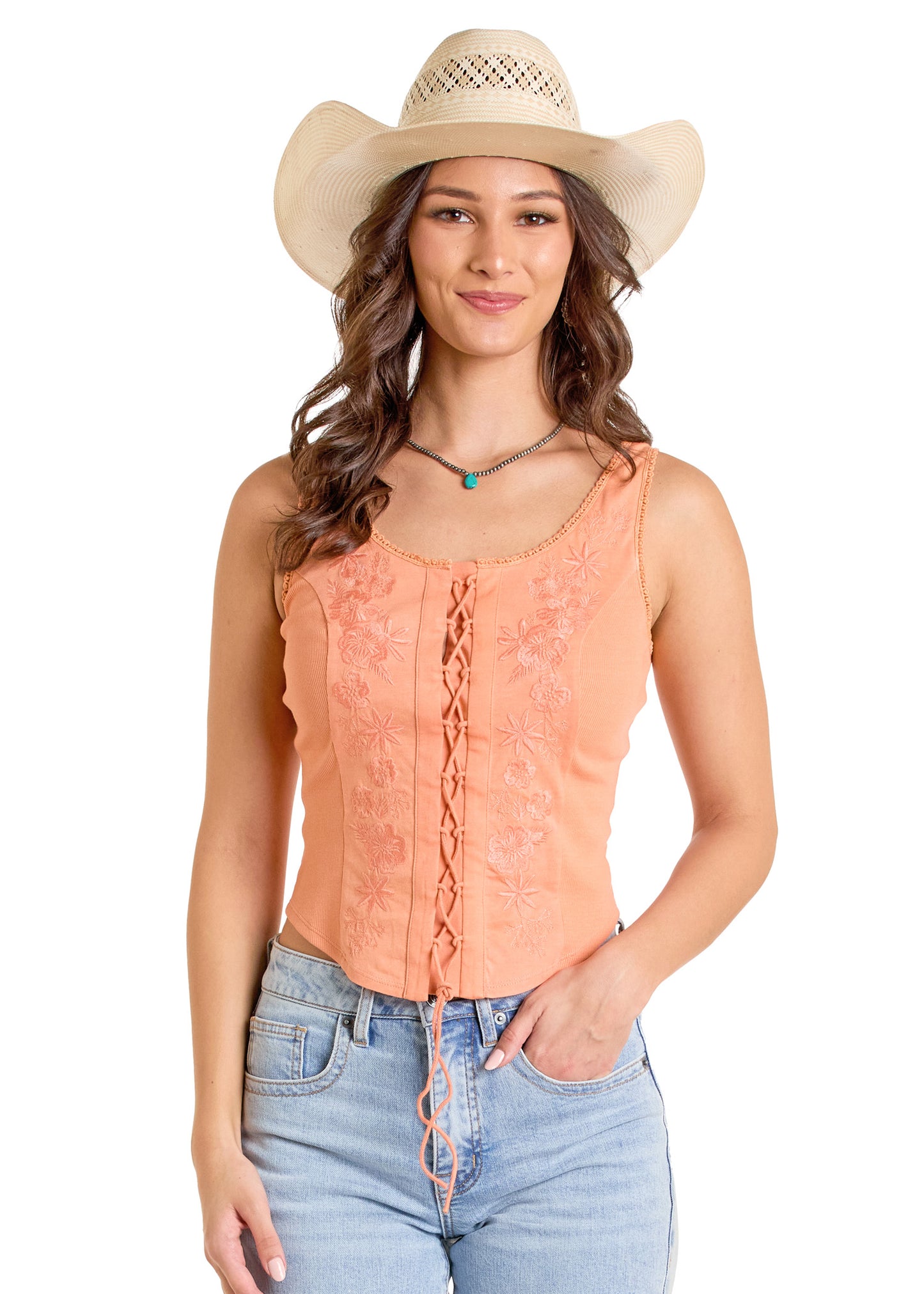 A woman wears the Rock & Roll Denim Embroidered Lace-Up Tank with floral details, a light brown cowboy hat, blue jeans, and a beaded necklace, smiling with one hand in her pocket against a white background.