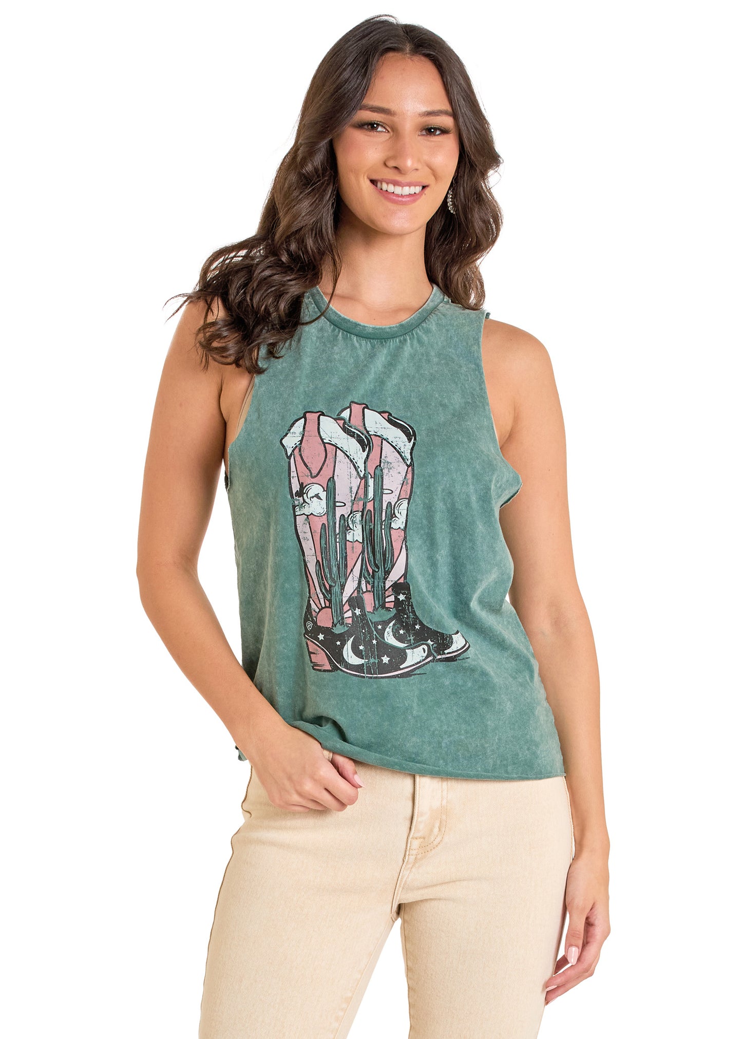 A woman with long brown hair wears a Rock & Roll Denim Desert Boots Graphic Tank Top—a green sleeveless top with cowboy boots, cacti, and mountains—paired with light beige pants, smiling as she stands against a white background.