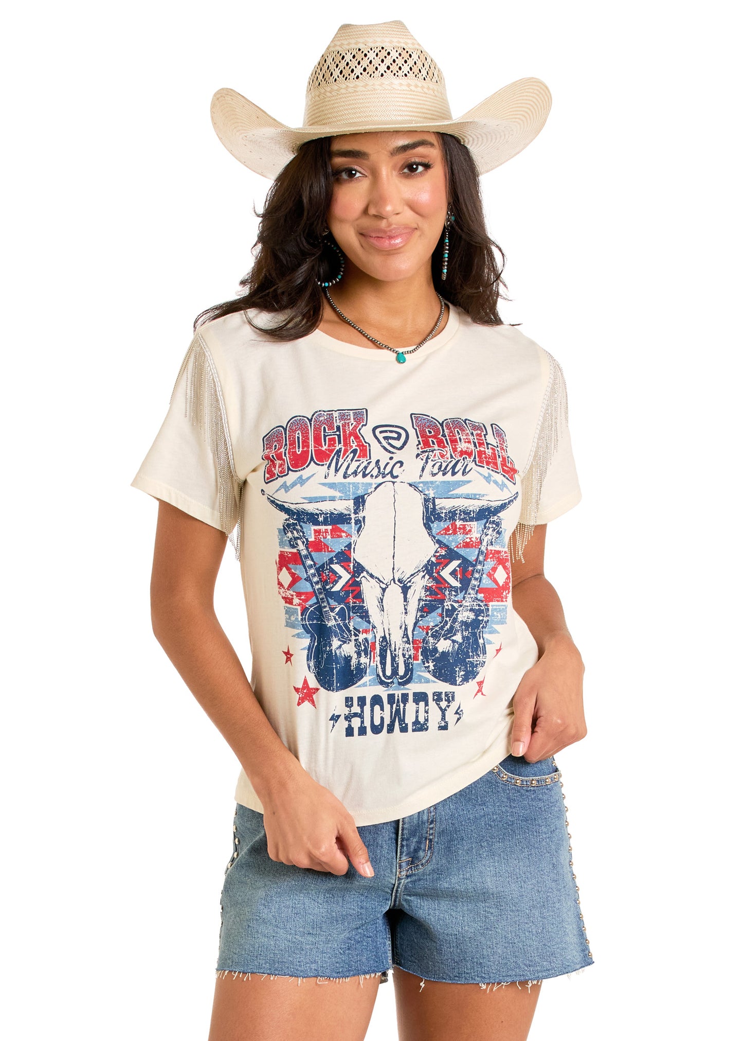 A woman in a cowboy hat, denim shorts, and a beige "Chain Fringe Rock & Roll Graphic Tee" by Rock & Roll Denim stands smiling with her hand in her pocket against a white background.
