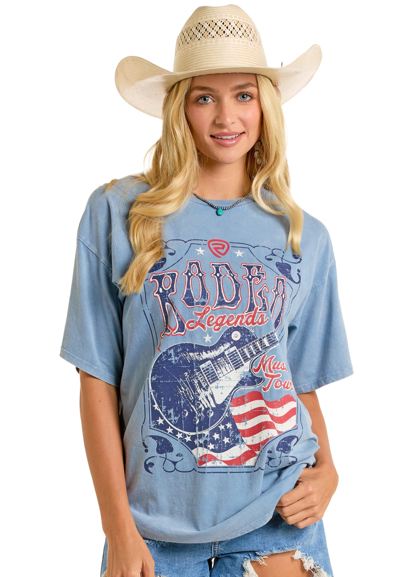 A young woman smiles in a straw cowboy hat and Rock & Roll Denim’s Rodeo Legends Oversized Tee, featuring a distressed guitar and American flag graphic, posed against a plain white background.