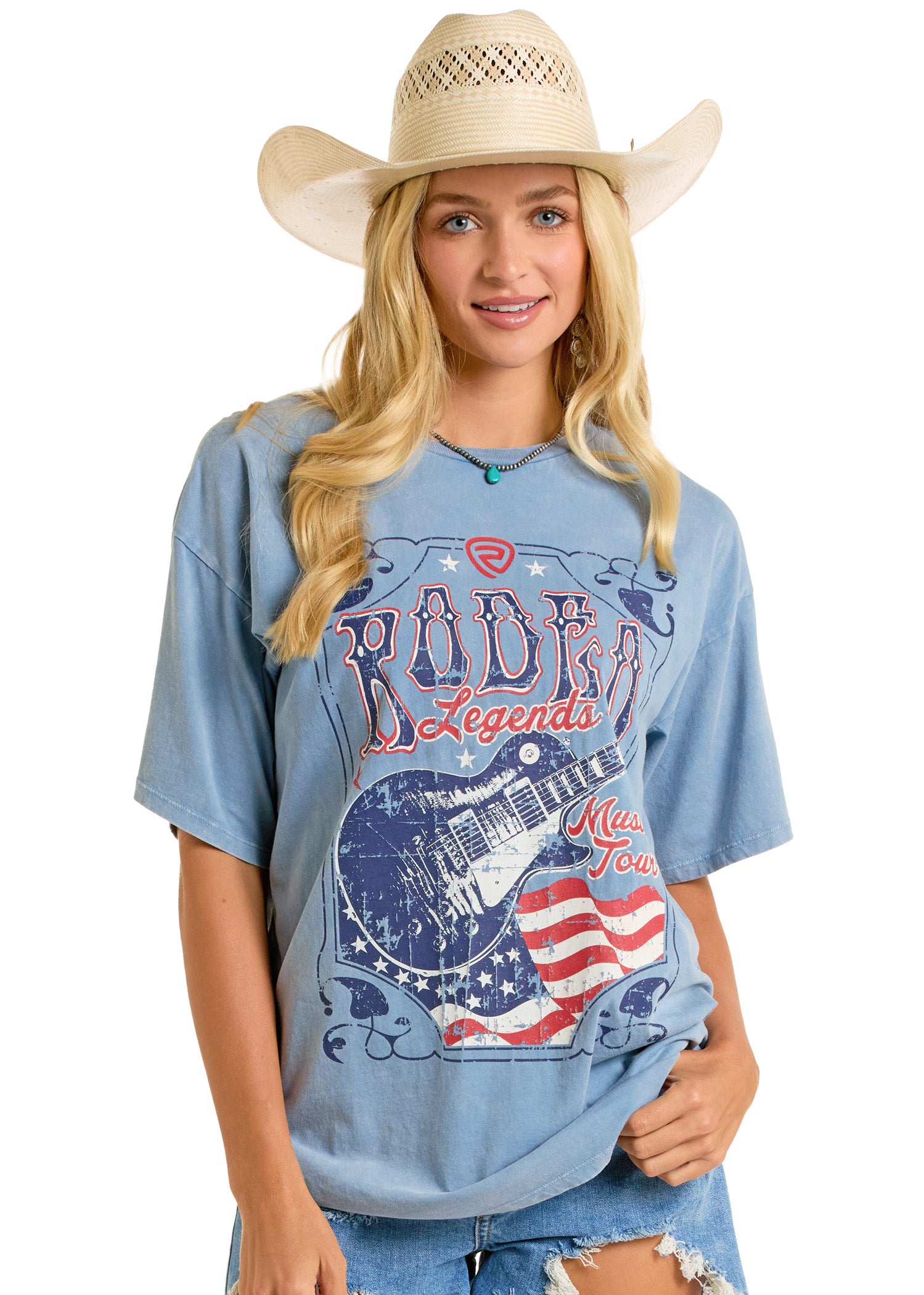 A young woman smiles in a straw cowboy hat and Rock & Roll Denim’s Rodeo Legends Oversized Tee, featuring a distressed guitar and American flag graphic, posed against a plain white background.