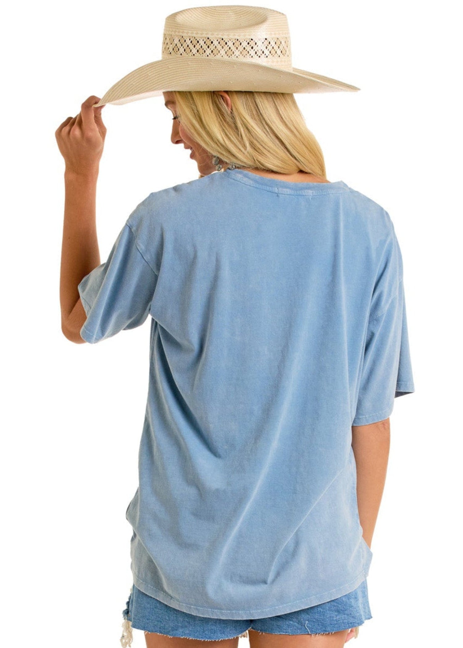 A woman with long blonde hair faces away, wearing a Rock & Roll Denim Rodeo Legends Oversized Tee, frayed denim shorts, and a straw hat held in one hand.
