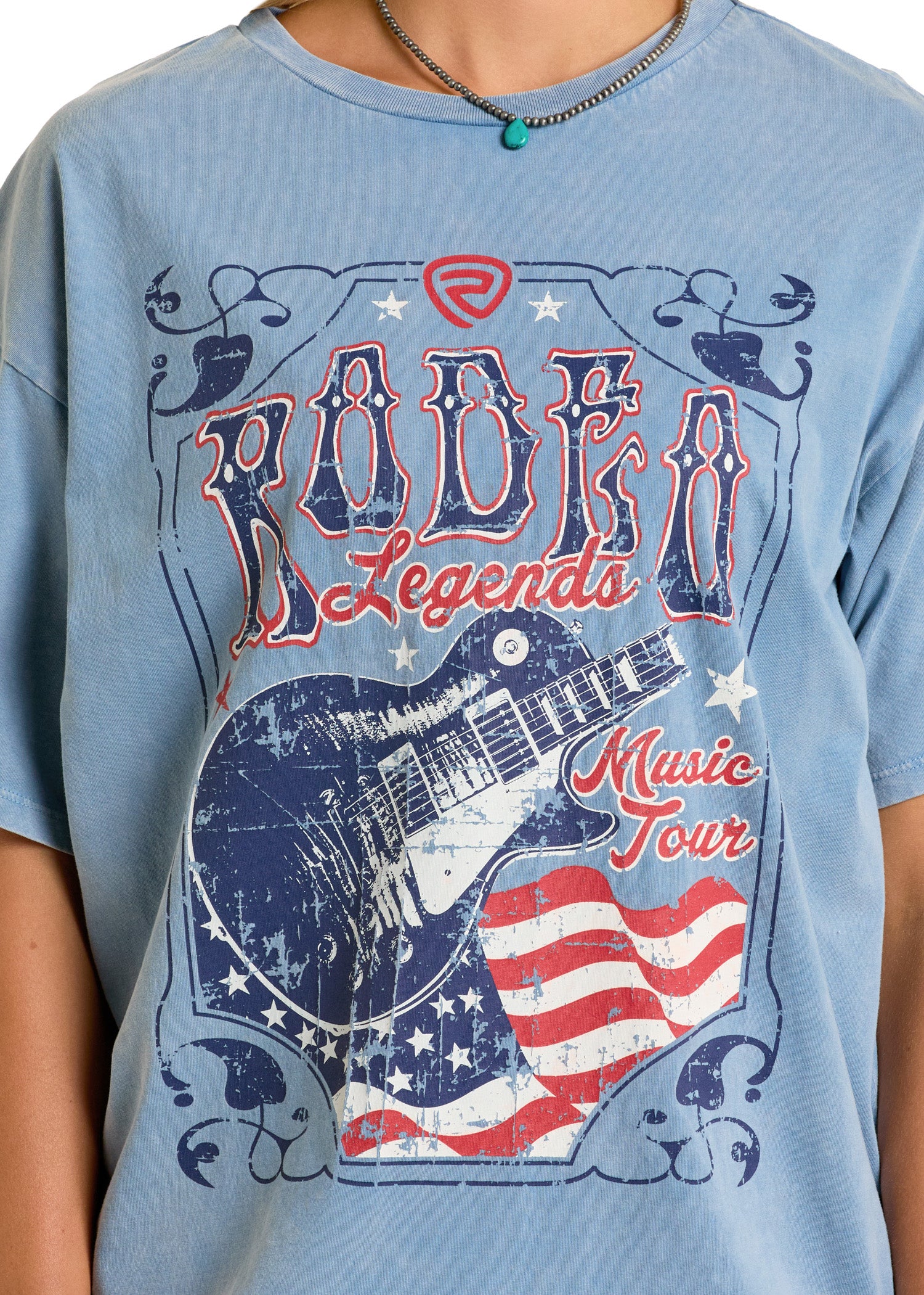 A person wears the Rock & Roll Denim Rodeo Legends Oversized Tee, showcasing a distressed guitar graphic with American flag details and bold "Rodeo Legends Music Tour" text in red, white, and blue.