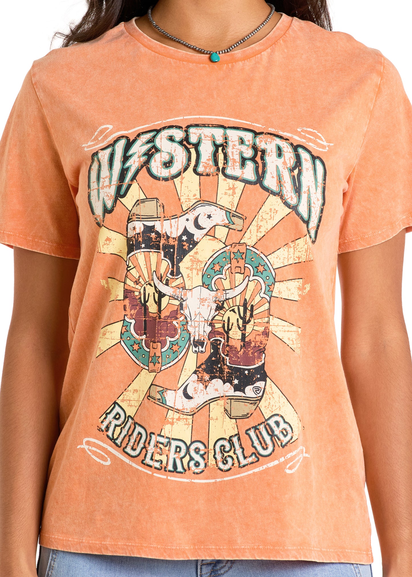 Someone wears the Rock & Roll Denim Western Riders Graphic Tee, featuring a steer skull, boot, cacti, and sunburst design, paired with a black necklace accented by a turquoise bead.