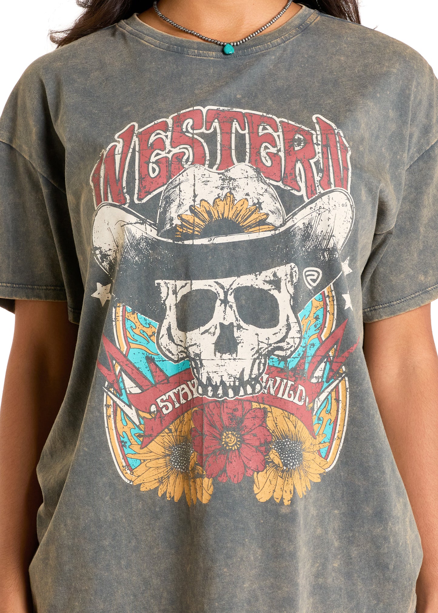 A person wears the Rock & Roll Denim Western Skull Oversized Tee—a faded gray, relaxed fit shirt featuring a distressed skull in a cowboy hat, sunflowers, bold “WESTERN” and “STAY WILD” text—styled with a turquoise necklace.