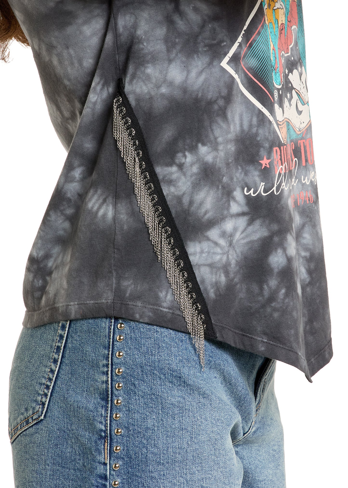 A person wears blue jeans with metal studs and a Rock & Roll Denim Chain Fringe Graphic Tee, which has decorative metal chain fringe along a diagonal seam and a colorful front graphic with text.