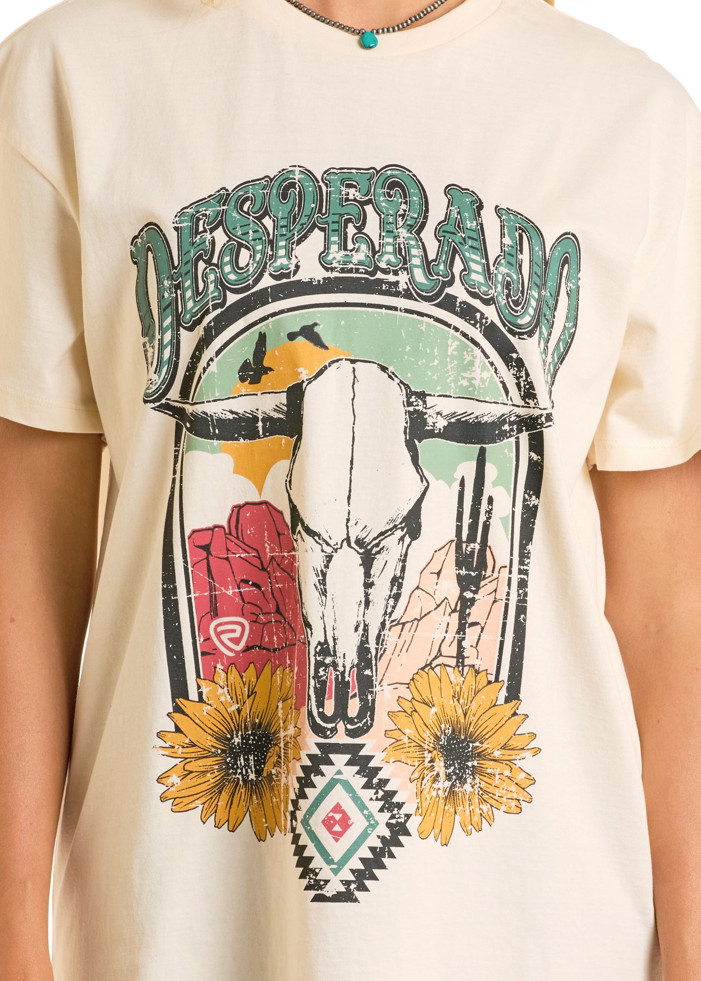 A person wearing the Sunflower Desert Oversized Tee by Rock & Roll Denim—a cream oversized tee with a distressed graphic of a cow skull, sunflowers, cacti, red rock cliffs, and bold "DESPERADO" lettering across the top.