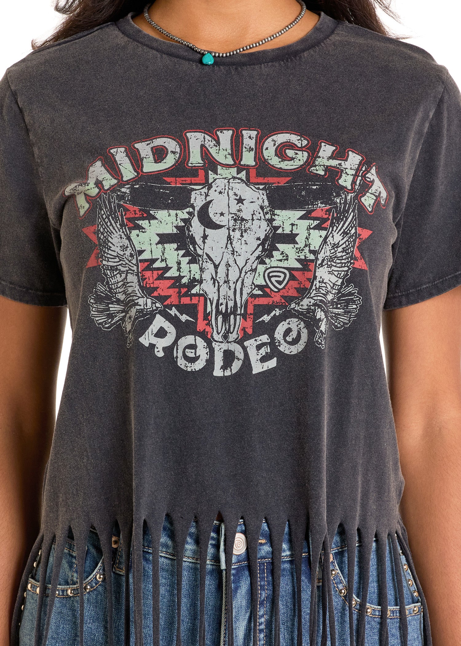 A close-up of a person wearing the Rock & Roll Denim Midnight Rodeo Fringe Tee, a black cropped tee with fringe, steer skull, eagles, and bold lettering. The look is styled with a beaded necklace for western flair.