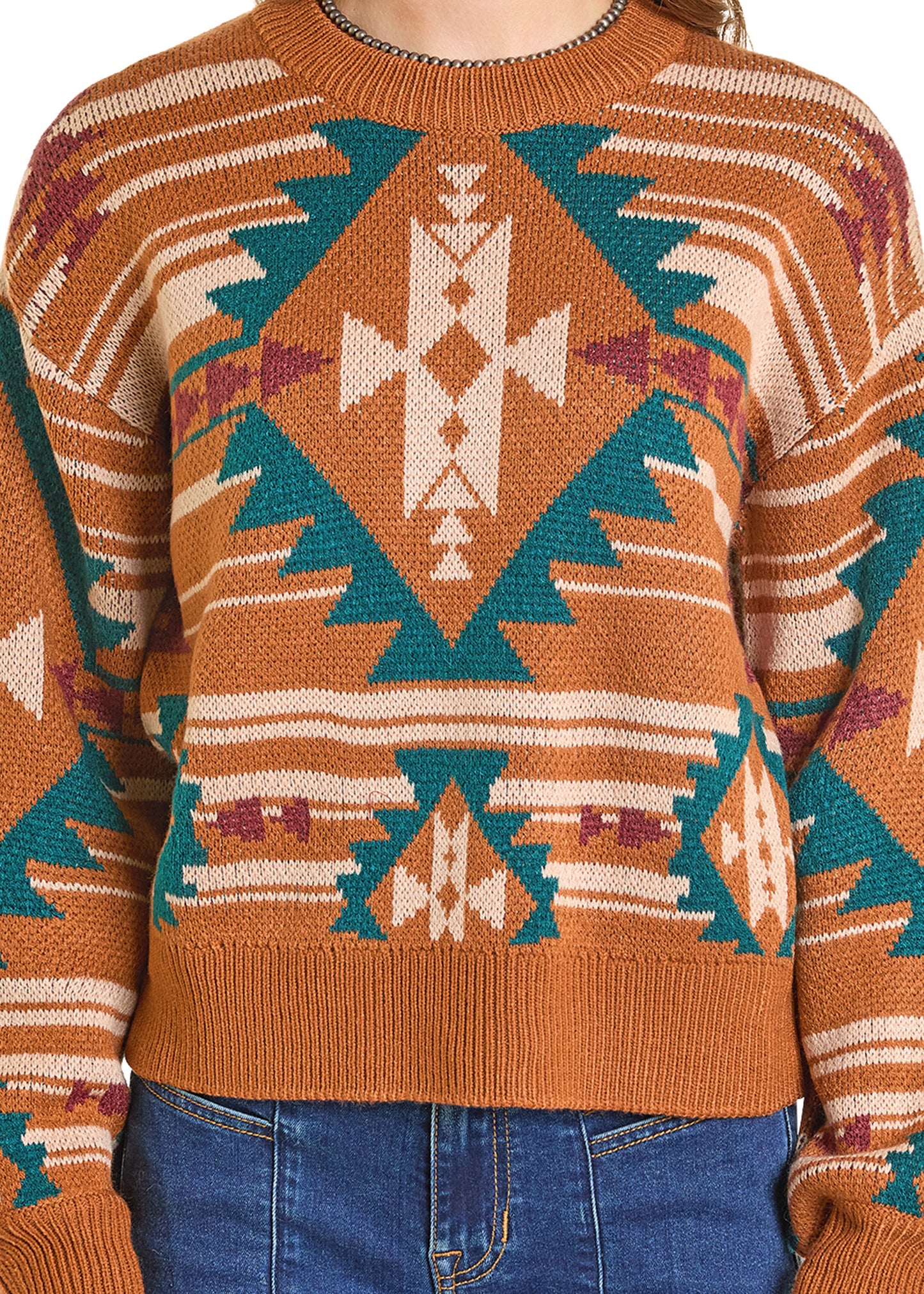 A person wears the Rock & Roll Denim Multicolor Aztec Knit Sweater featuring teal, white, and maroon Southwestern geometric patterns with blue jeans. The image is cropped at the neck and hips.
