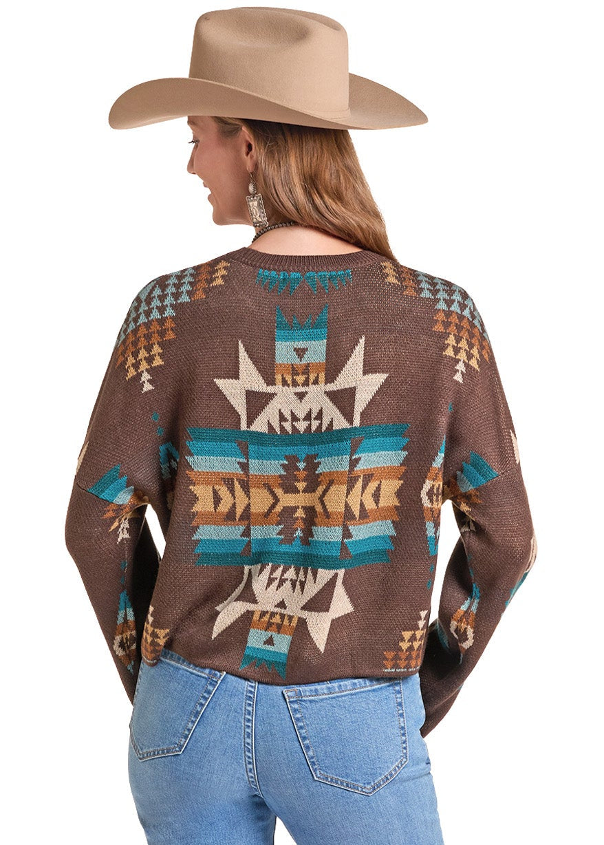 A woman wearing a brown long-sleeve knit sweater with a colorful Southwestern geometric pattern, paired with light blue jeans and a tan cowboy hat.