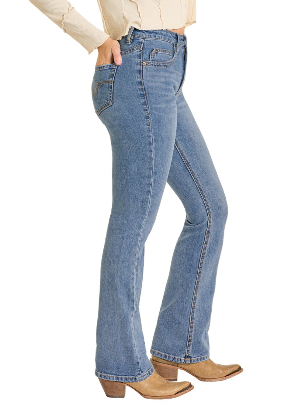 A person wears Rock & Roll Denim High Rise Yoke Stitch Bootcut Jeans, a cream long sleeve shirt, and tan heeled boots, standing with one hand in a pocket against a white background.