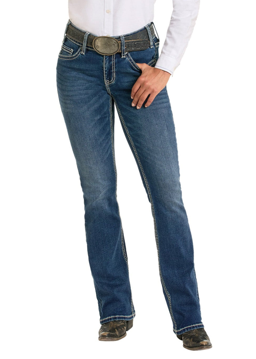 A person wearing a white shirt, Rock & Roll Denim Mid Rise Cowhide Stitch Bootcut Riding Jeans, a wide Western belt with a large buckle, and worn black boots stands with one hand in their pocket.