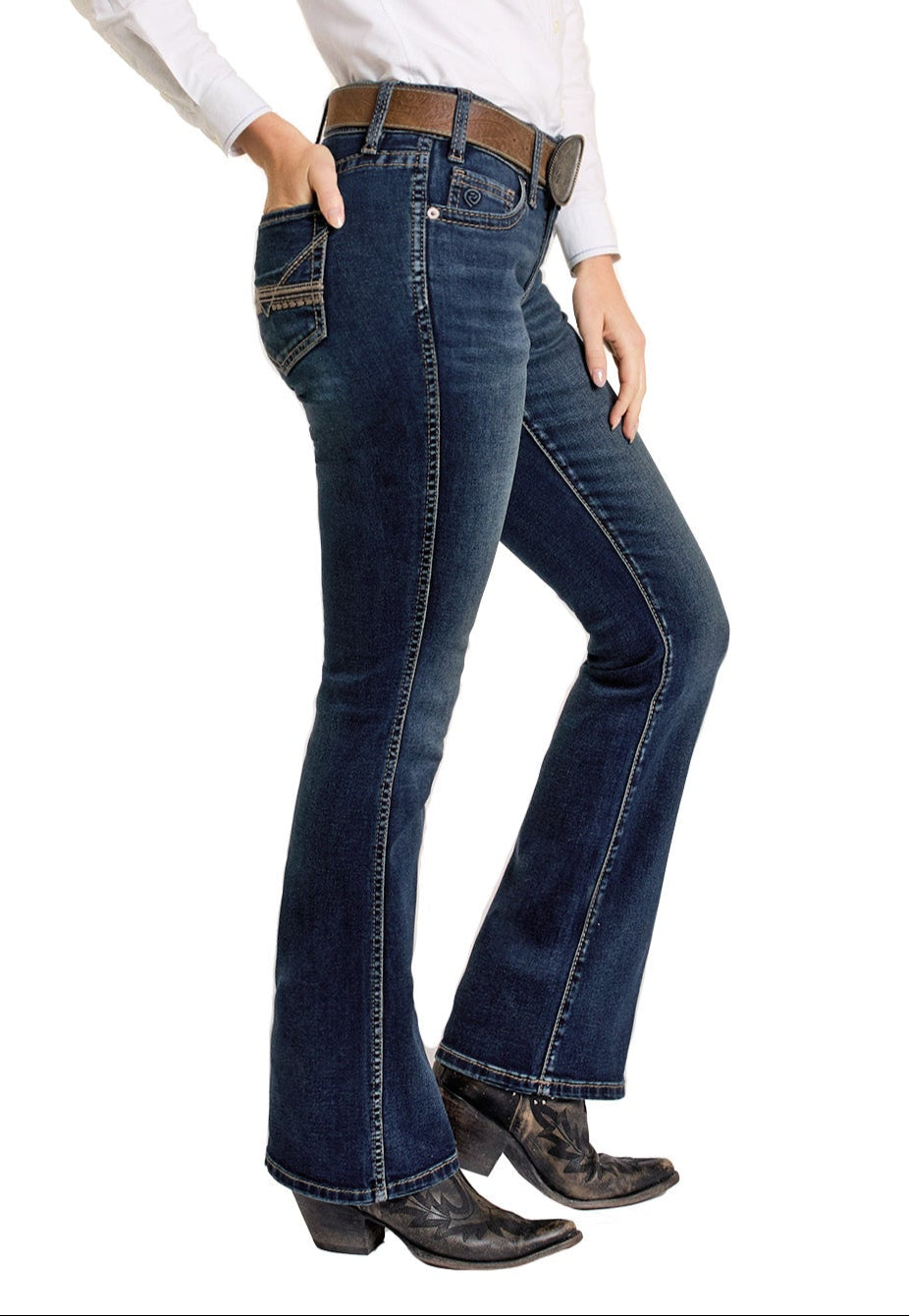 A person wearing Rock & Roll Denim Mid Rise Classic Stitch Bootcut Riding Jeans, a white shirt, a brown belt with a large buckle, and black cowboy boots stands with one hand in their pocket against a white background.