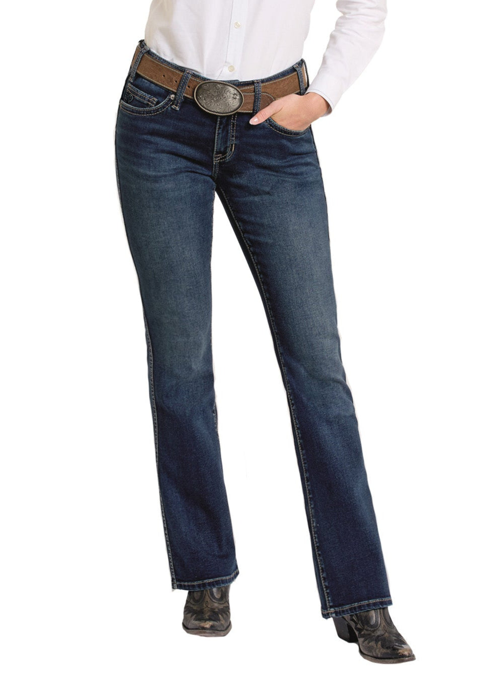 A person wears Rock & Roll Denim Mid Rise Classic Stitch Bootcut Riding Jeans in dark blue, a brown leather belt with a large oval buckle, a white shirt, and black cowboy boots, with one hand in their pocket.