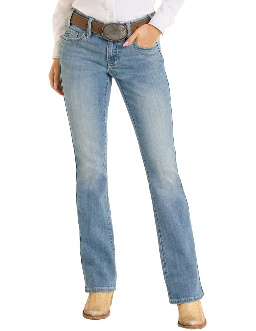 A person wearing light blue Rock & Roll Denim Mid Rise Cross Stitch Bootcut Riding Jeans, a white shirt, a brown belt with a large buckle, and tan cowboy boots stands with one hand in their pocket against a white background.