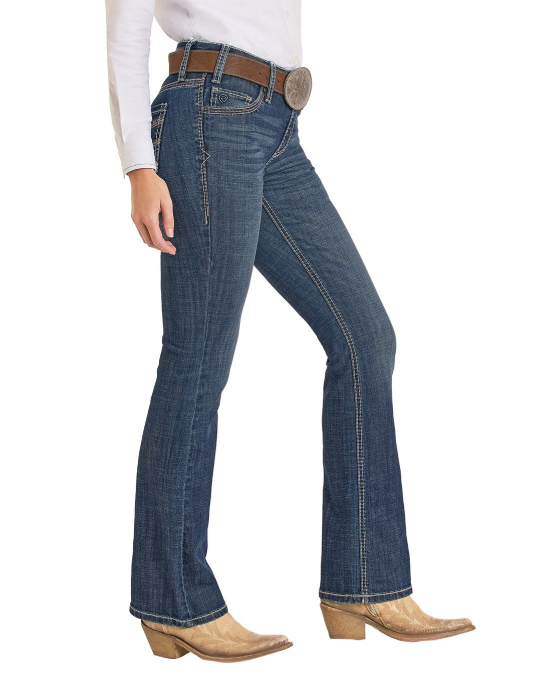 A person wearing Rock & Roll Denim's Mid Rise Cross Stitch Bootcut Riding Jeans, a white tucked-in shirt, brown belt with a large buckle, and tan cowboy boots stands in profile against a white background.