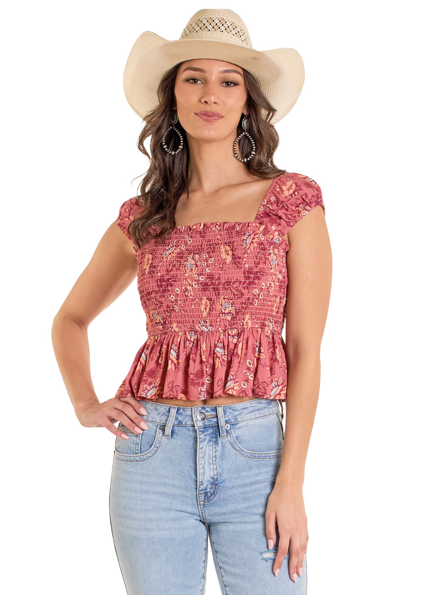 A woman in a white cowboy hat, hoop earrings, and the Rock & Roll Denim Smocked Floral Top with a flowy bodice and elastic cap sleeves, paired with light blue jeans, stands with one hand on her hip and smiles slightly.