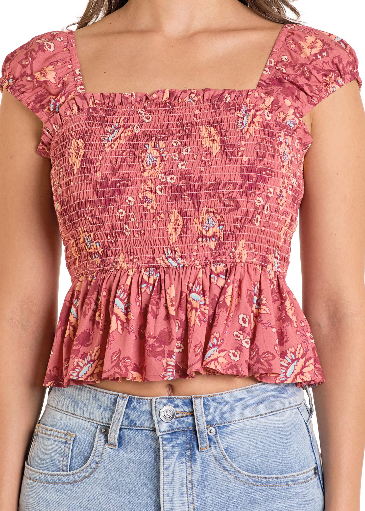 A woman wears the Rock & Roll Denim Smocked Floral Top with elastic cap sleeves, a pink floral print, ruffled hem, square neckline, and fitted ruched bodice, paired with light blue high-waisted jeans.