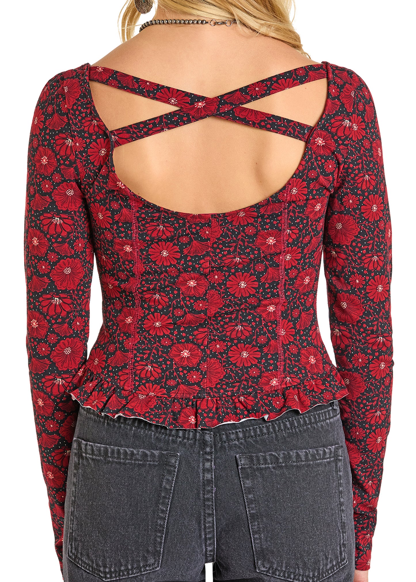 A person wears the Rock & Roll Denim Floral Jersey Top, featuring a red floral print, crisscross open back, and ruffled hem, paired with dark high-waisted jeans. The rear view highlights the trendy cross back and printed pattern.