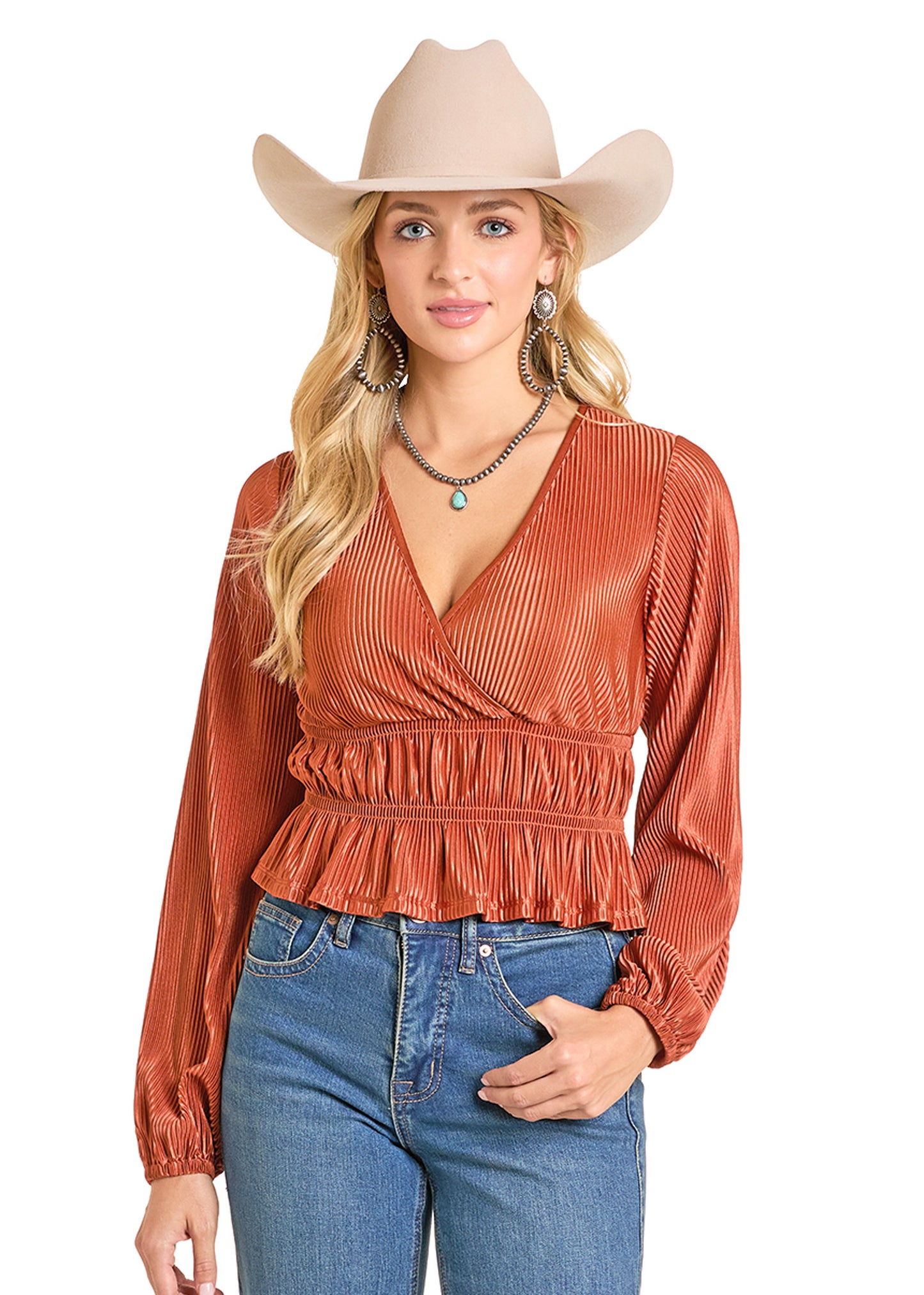 A woman in a rust-colored Rock & Roll Denim Velvet Plisse Blouse with wrap v-neckline and ruffled hem, blue jeans, hoop earrings, and layered necklaces poses against a white background, wearing a beige cowboy hat.