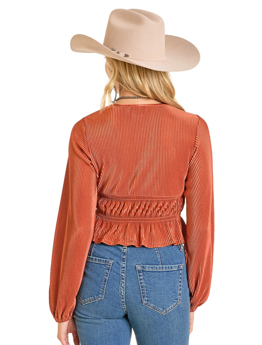 A woman with long blonde hair, facing away from the camera, wears a beige wide-brimmed hat, blue jeans, and a Rock & Roll Denim Velvet Plisse Blouse with an orange hue and ruffled hem.