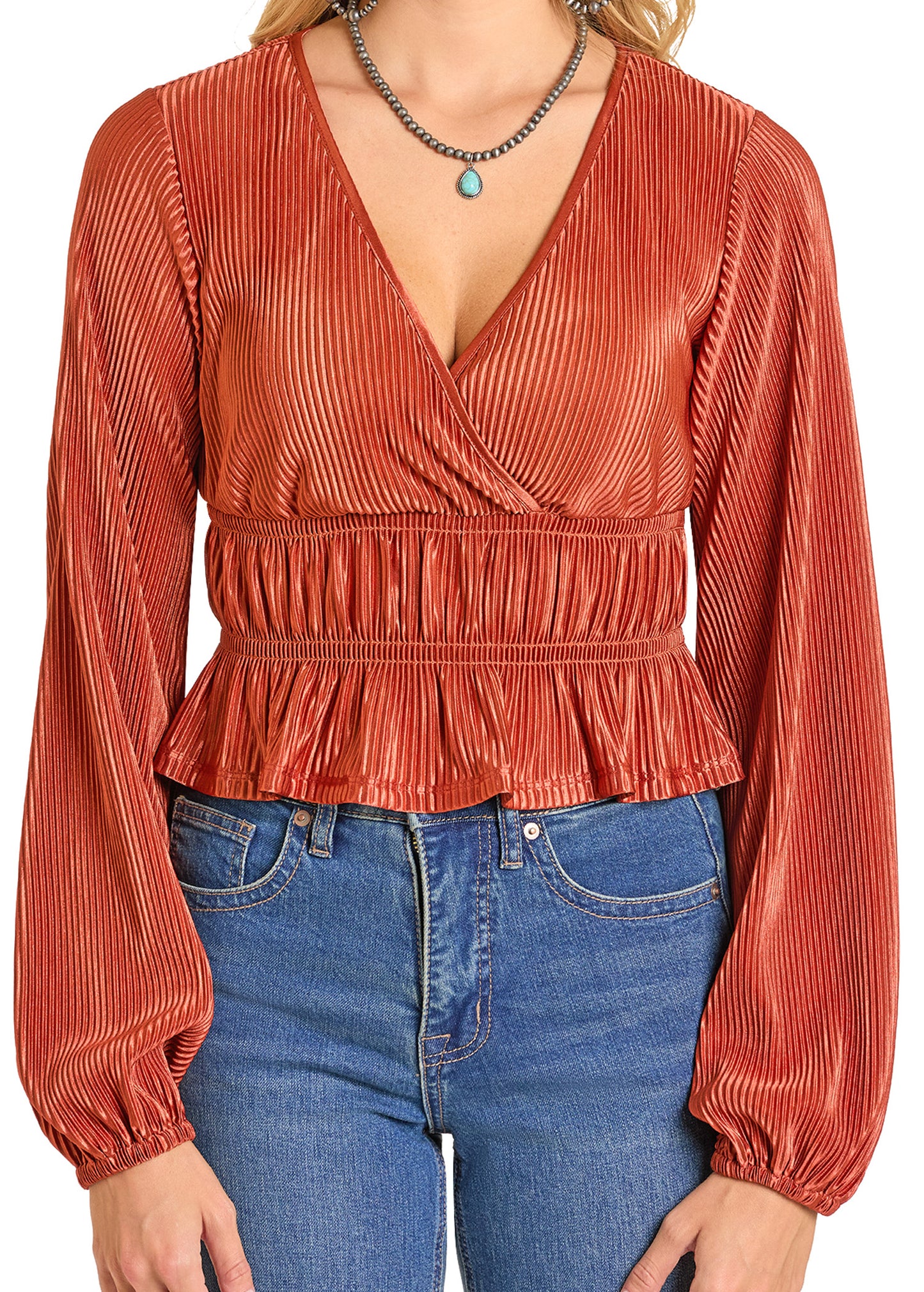 A woman wears a rust-colored Rock & Roll Denim Velvet Plisse Blouse with a wrap v-neckline, ruffled hem, and high-waisted blue jeans, accessorized with a turquoise pendant necklace.