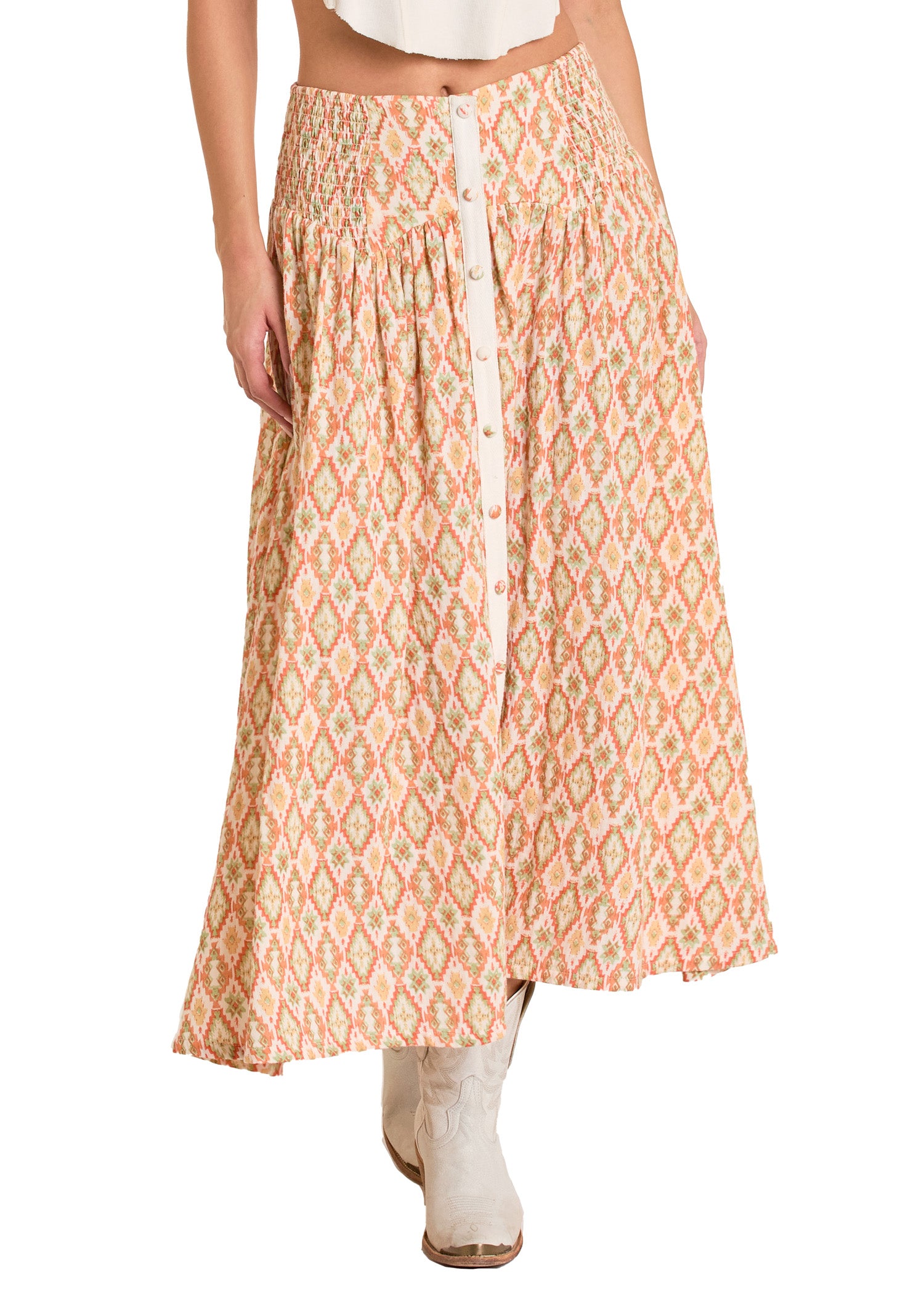 A woman wears the Rock & Roll Denim Aztec Print Maxi Skirt in peach and cream, featuring a smocked elastic waist and front button row, styled with white boots. The image shows her from the waist down.