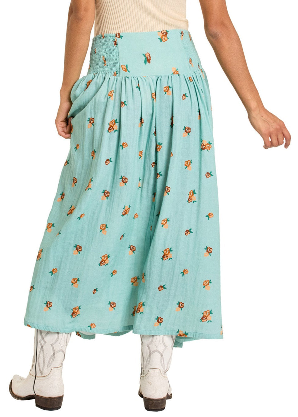 A person wearing a light beige top, Rock & Roll Denim Floral Print Maxi Skirt - XXL in turquoise with orange floral pattern and smocked waist, paired with white cowboy boots, shown from the back.