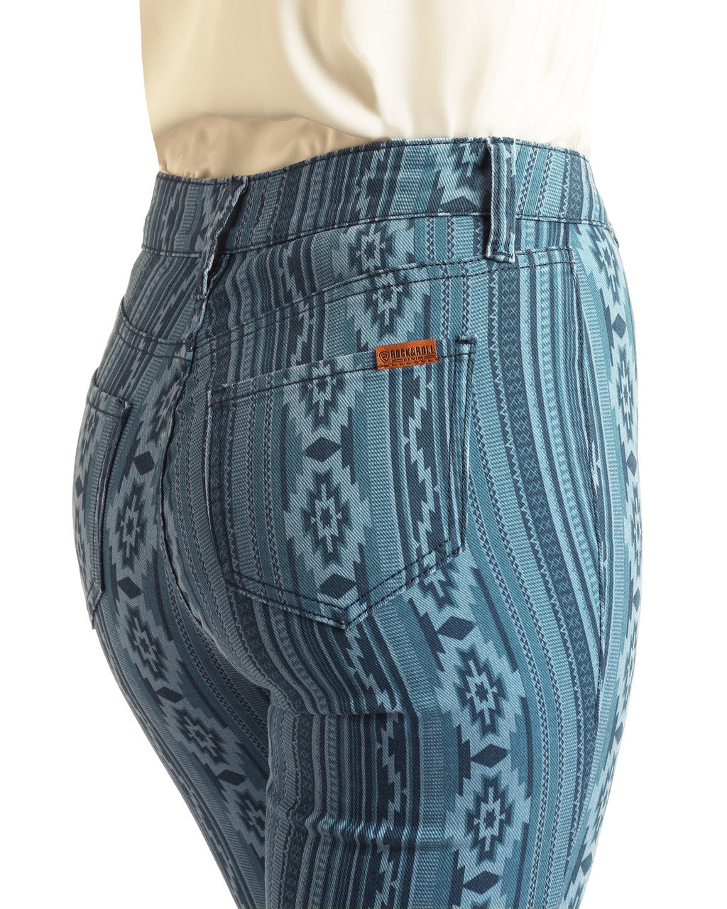 A person wearing Rock & Roll Denim Diamonds & Stripes Aztec High Rise Flare Jeans with a blue geometric pattern and a beige top, shown from the waist down and viewed from behind; jeans feature a brown brand tag on the back pocket.